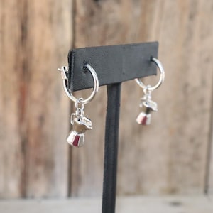 May include: A pair of silver hoop earrings with small, detailed coffee maker charms. The charms have a red accent. The earrings are displayed on a black stand against a blurred wooden background. The earrings are a jewelry item.