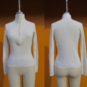 May include: A white ribbed sweater with a zipper at the neck. The sweater has long sleeves and a fitted silhouette.
