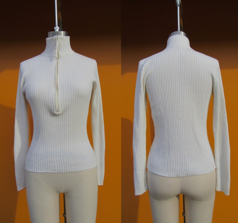 May include: A white ribbed sweater with a zipper at the neck. The sweater has long sleeves and a fitted silhouette.