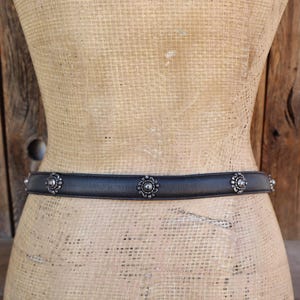 May include: A black leather belt with silver floral detailing. The belt is shown on a mannequin against a wooden backdrop. The floral accents are evenly spaced along the belt's length, adding a decorative touch to the accessory.