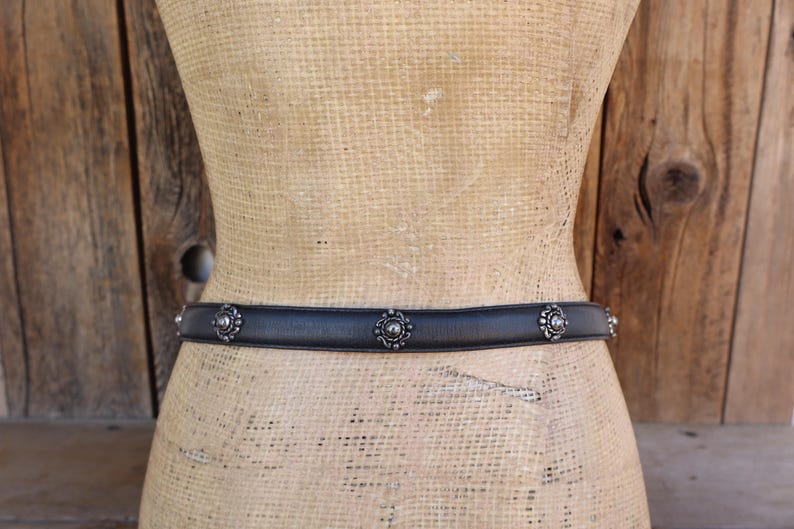 May include: A black leather belt with silver floral detailing. The belt is shown on a mannequin against a wooden backdrop. The floral accents are evenly spaced along the belt's length, adding a decorative touch to the accessory.
