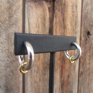 May include: A pair of silver hoop earrings with a gold accent on a black display stand. The earrings are made of two hoops, one larger than the other, with the smaller hoop sitting inside the larger hoop.