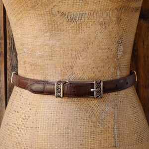 May include: A brown leather belt with a silver buckle and decorative accents. The belt is displayed on a mannequin and has a textured surface. The buckle has a rectangular shape with a silver finish.