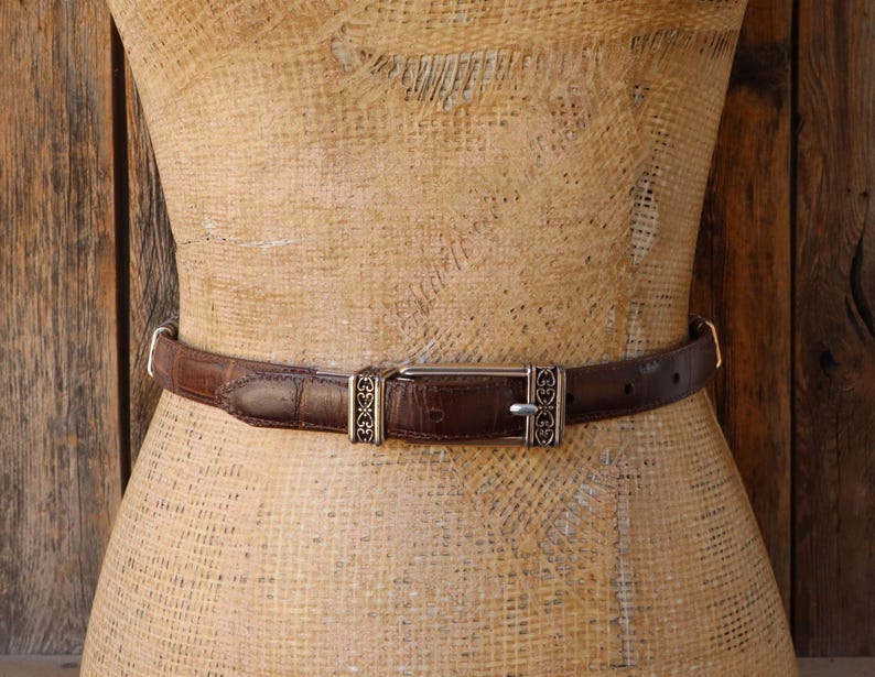 May include: A brown leather belt with a silver buckle and decorative accents. The belt is displayed on a mannequin and has a textured surface. The buckle has a rectangular shape with a silver finish.