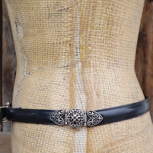 May include: A black leather belt with a decorative silver-tone buckle. The buckle features an ornate design with a central floral element and heart-shaped accents. The belt is displayed on a mannequin against a rustic wooden background.