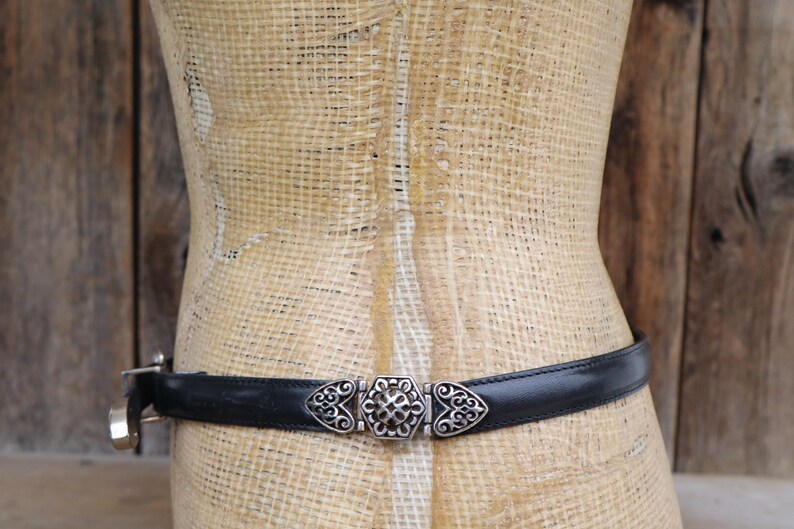 May include: A black leather belt with a decorative silver-tone buckle. The buckle features an ornate design with a central floral element and heart-shaped accents. The belt is displayed on a mannequin against a rustic wooden background.