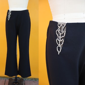 Puede incluir: Pantalones negros acampanados con un diseño decorativo de pedrería en el lateral. Los pantalones se exhiben en un maniquí sobre un fondo amarillo. El diseño de pedrería es una serie de formas de hojas conectadas.