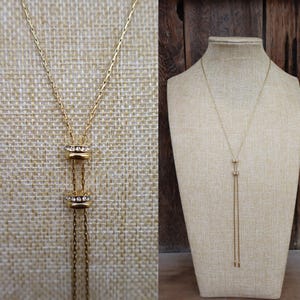 May include: Gold-toned necklace with a sliding pendant design. The necklace features two stacked, cylindrical elements adorned with clear crystals. The chain extends into a delicate, dangling strand. The necklace is displayed on a beige mannequin and a textured background.