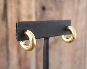 May include: A pair of gold-colored, chunky hoop earrings. The earrings are displayed on a black earring stand against a blurred wooden background. The hoops have a smooth, polished finish.