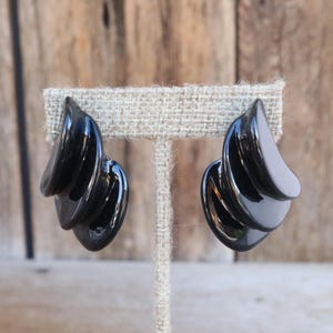 Op de afbeelding: Een paar zwarte, glanzende oorbellen met een sculpturaal ontwerp. Elke oorbel heeft drie gebogen, gelaagde elementen die een golfvorm creëren. De oorbellen worden gepresenteerd op een neutrale, getextureerde ondergrond.