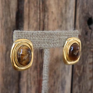 Könnte beinhalten: Ein Paar goldfarbene Clip-Ohrringe mit ovalen, braunen und bernsteinfarbenen Steinen. Die Ohrringe haben eine rechteckige Form mit abgerundeten Kanten und werden auf einem neutralen Stoff präsentiert.