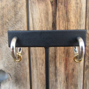 May include: A pair of silver hoop earrings with a gold accent. The earrings are displayed on a black stand.