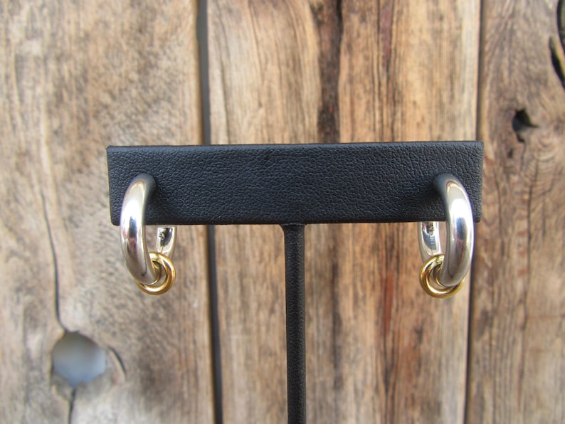 May include: A pair of silver hoop earrings with a gold accent. The earrings are displayed on a black stand.