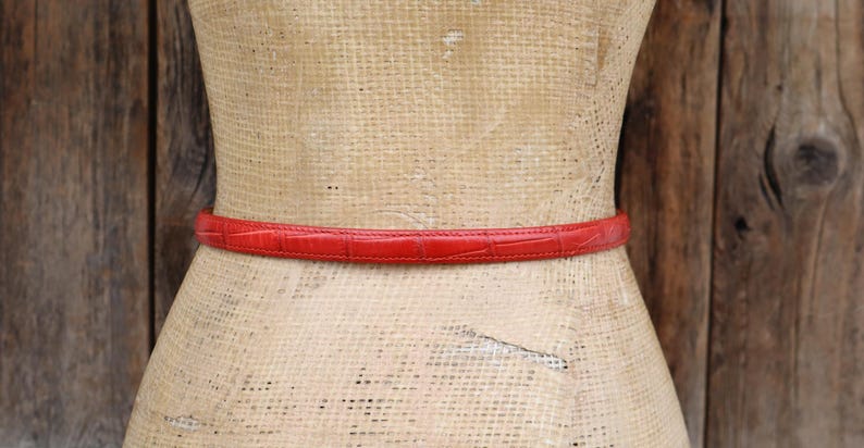 Puede incluir: Un cintur&oacute;n rojo texturizado envuelve la cintura de un maniqu&iacute;. El cintur&oacute;n es un material delgado similar al cuero con un patr&oacute;n repetitivo. El maniqu&iacute; est&aacute; hecho de un material tejido de color claro, y el fondo es una pared de madera.