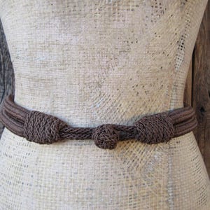 Può includere: Una cintura in corda intrecciata marrone con un design annodato. La cintura è legata attorno a un manichino rivestito in tessuto.