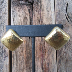 May include: A pair of gold-toned hammered metal earrings in a diamond shape. The earrings are displayed on a black stand against a wooden background.