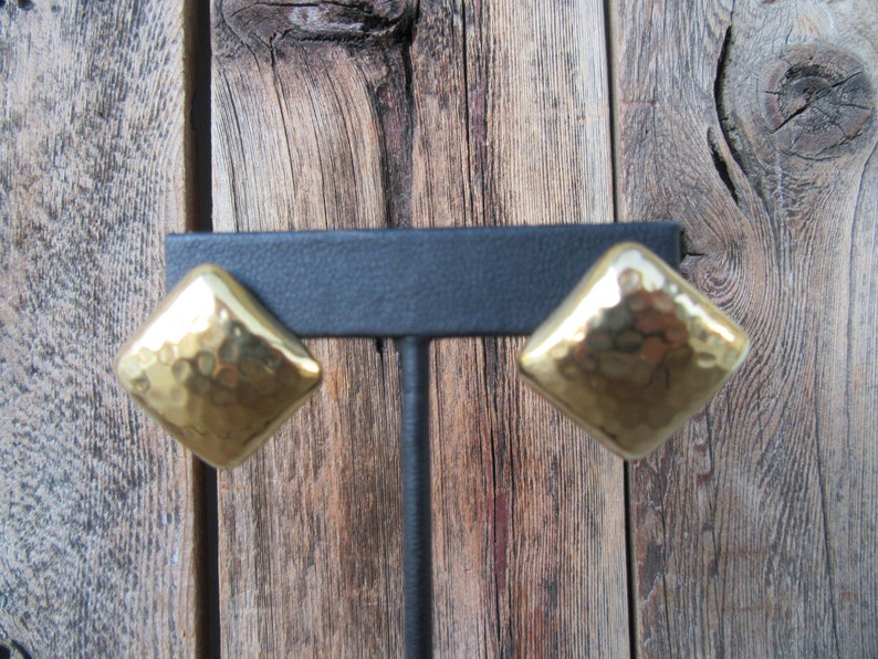 May include: A pair of gold-toned hammered metal earrings in a diamond shape. The earrings are displayed on a black stand against a wooden background.