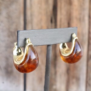 May include: A pair of vintage hoop earrings. The earrings feature a teardrop shape with a gold-tone metal accent and a translucent amber-colored resin. The earrings are displayed on a gray stand against a wooden background.