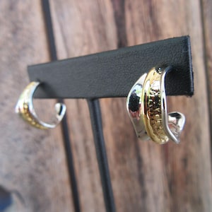 May include: A pair of silver and gold hoop earrings. The earrings are made of two different metals, with a silver band on the outside and a gold band on the inside. The earrings are simple and elegant.