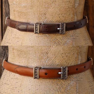 May include: Two leather belts, one brown and one tan, displayed on a mannequin. Both belts have silver-colored buckles with decorative filigree designs. The brown belt has a textured surface, while the tan belt is smooth.