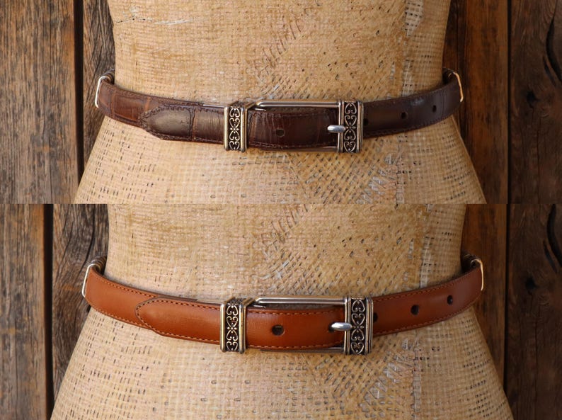 May include: Two leather belts, one brown and one tan, displayed on a mannequin. Both belts have silver-colored buckles with decorative filigree designs. The brown belt has a textured surface, while the tan belt is smooth.