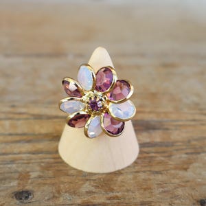 May include: A gold-toned flower ring with purple and white gemstones. The ring is set on a wooden display cone. The flower design features a central purple stone surrounded by petals of varying shades of purple and white.
