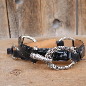 May include: Black belt with silver-tone metal hardware. The belt features a glossy, textured finish and decorative silver rings and a clasp. The belt is lying on a wooden surface.