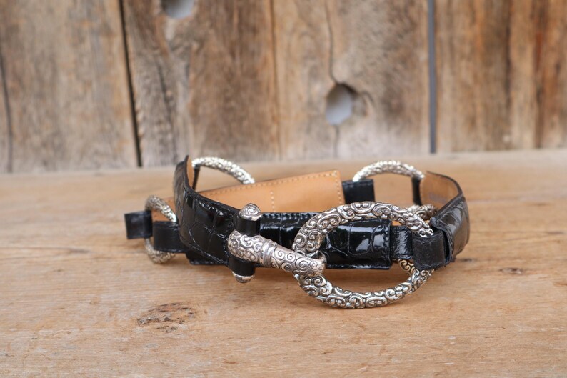May include: Black belt with silver-tone metal hardware. The belt features a glossy, textured finish and decorative silver rings and a clasp. The belt is lying on a wooden surface.