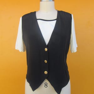 Peut inclure: Un gilet sans manches noir avec des boutons dor&eacute;s. Le gilet a un col en V et une ourlet pointu.
