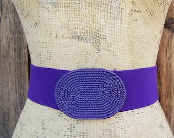 May include: A purple elastic belt with a large oval-shaped buckle. The buckle is made of woven purple rope.