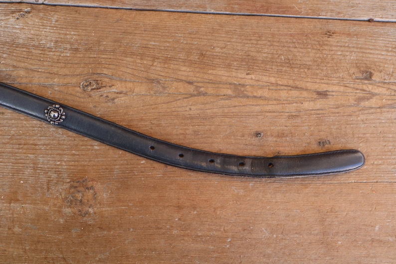 May include: A black leather belt with a decorative silver buckle. The belt has several holes for adjustment and shows signs of wear. The background is a wooden surface.