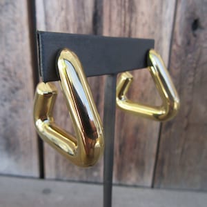 May include: A pair of gold-tone hoop earrings with a triangular shape. The earrings are displayed on a black stand.