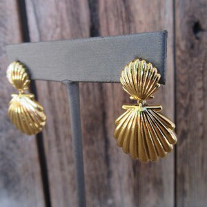 May include: Gold tone earrings with a double seashell design. The earrings are on a gray display stand.