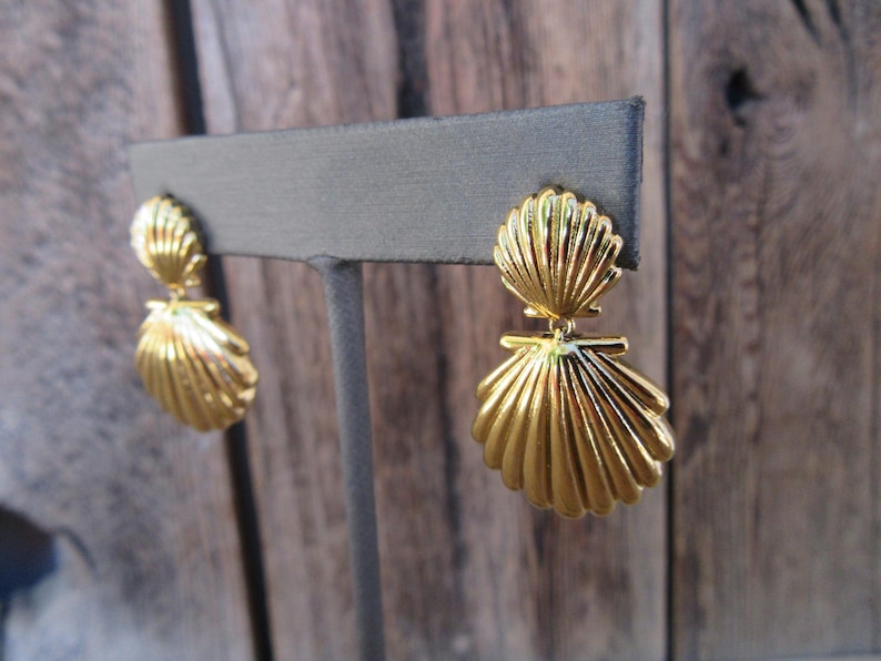 May include: Gold tone earrings with a double seashell design. The earrings are on a gray display stand.