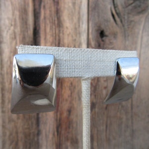 May include: A pair of polished silver-toned square earrings with a curved design. The earrings are displayed on a neutral-colored linen earring stand against a rustic wooden background. The earrings have a reflective surface.