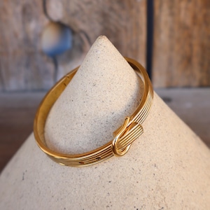 May include: A gold-colored bracelet with a clasp closure. The bracelet has a series of parallel lines and is displayed on a light-colored, cone-shaped object. The background is a wooden surface.