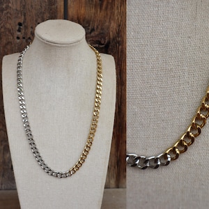 May include: A two-tone curb chain necklace. One half of the necklace is silver-colored, and the other half is gold-colored. The necklace is displayed on a mannequin with a beige fabric covering.