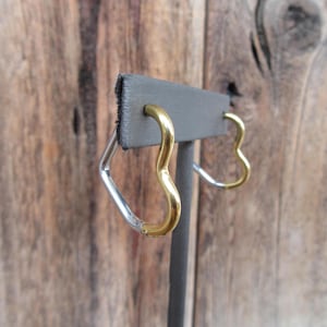 May include: A pair of heart-shaped earrings with a gold and silver finish. The earrings are displayed on a black stand.
