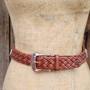 May include: A brown leather belt with a woven design is fastened around a mannequin. The belt has a silver-coloured buckle and silver accents. The mannequin is on a wooden background.