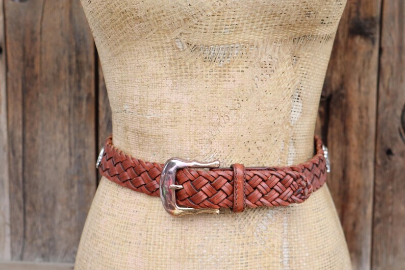 May include: A brown leather belt with a woven design is fastened around a mannequin. The belt has a silver-coloured buckle and silver accents. The mannequin is on a wooden background.