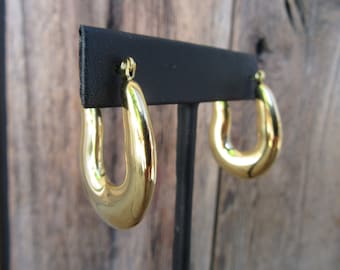 Pode incluir: Um par de brincos de argola dourados em forma de U. Os brincos são exibidos em um suporte preto contra um fundo de madeira desfocado. Os brincos têm uma superfície lisa e polida, refletindo a luz.