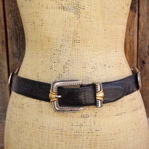 May include: A black leather belt with a silver and gold buckle. The belt is worn on a mannequin. The buckle has a rectangular shape with a silver frame and gold accents. The belt is approximately 1.5 inches wide.