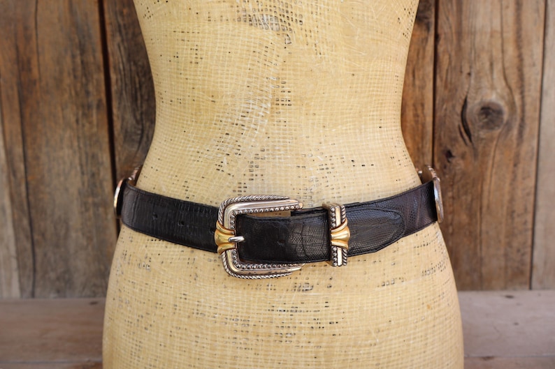 May include: A black leather belt with a silver and gold buckle. The belt is worn on a mannequin. The buckle has a rectangular shape with a silver frame and gold accents. The belt is approximately 1.5 inches wide.