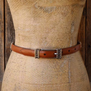 May include: A brown leather belt with a silver buckle and decorative accents. The belt is displayed on a mannequin. The belt has a classic design and is suitable for various outfits.