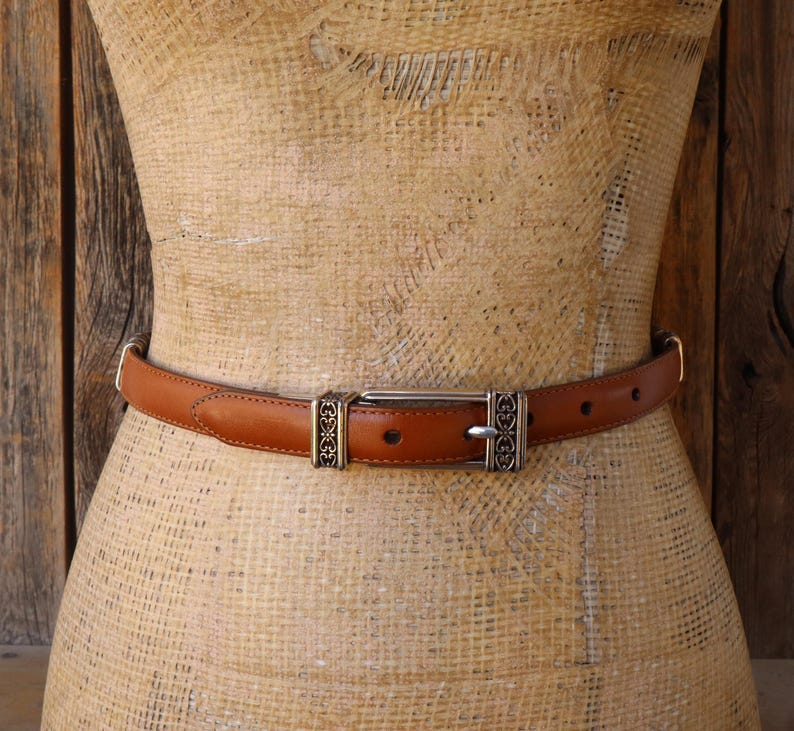 May include: A brown leather belt with a silver buckle and decorative accents. The belt is displayed on a mannequin. The belt has a classic design and is suitable for various outfits.