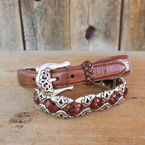 May include: Two brown leather belts with silver buckles and accents. The top belt has a classic buckle and a braided leather section. The bottom belt features a decorative silver and braided leather design. The belts are displayed on a wooden surface.