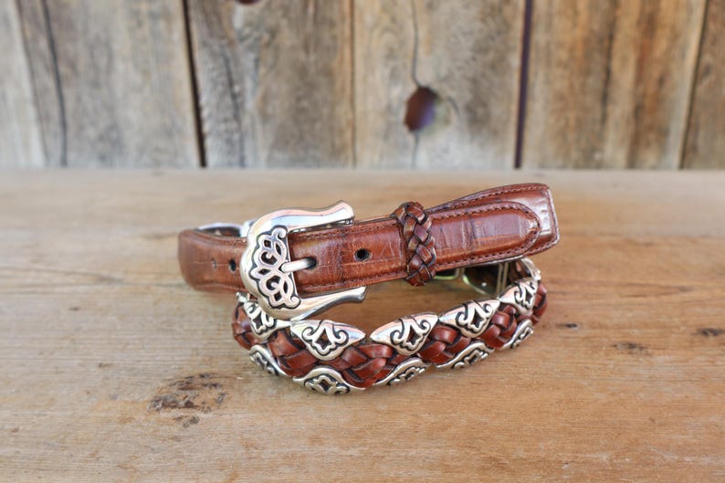 May include: Two brown leather belts with silver buckles and accents. The top belt has a classic buckle and a braided leather section. The bottom belt features a decorative silver and braided leather design. The belts are displayed on a wooden surface.
