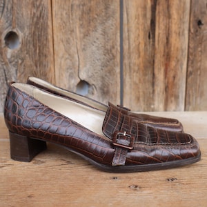 Puede incluir: Un par de mocasines de cuero marrón con textura de cocodrilo. Los zapatos tienen punta cuadrada, tacón pequeño y una correa con hebilla en la parte superior. Los zapatos se exhiben sobre una superficie de madera.