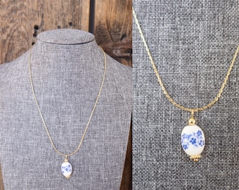 Könnte beinhalten: Eine goldfarbene Halskette mit einem weißen, ovalen Anhänger. Der Anhänger ist mit blauen Blumenmustern verziert. Die Halskette wird auf einer grauen Mannequin-Büste vor einem Holzgrund gezeigt.