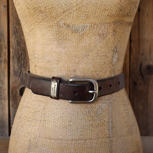 May include: A brown leather belt with a silver-colored buckle and a small metal tag that reads "Fossil 1954". The belt is worn around a mannequin. The belt is approximately 1.25 inches wide.